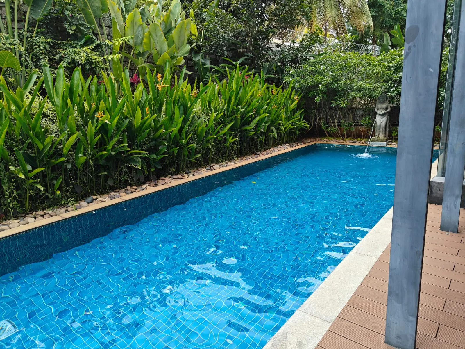 A tiled swimming pool framed by tropical greenery beside a contemporary Yelahanka villa